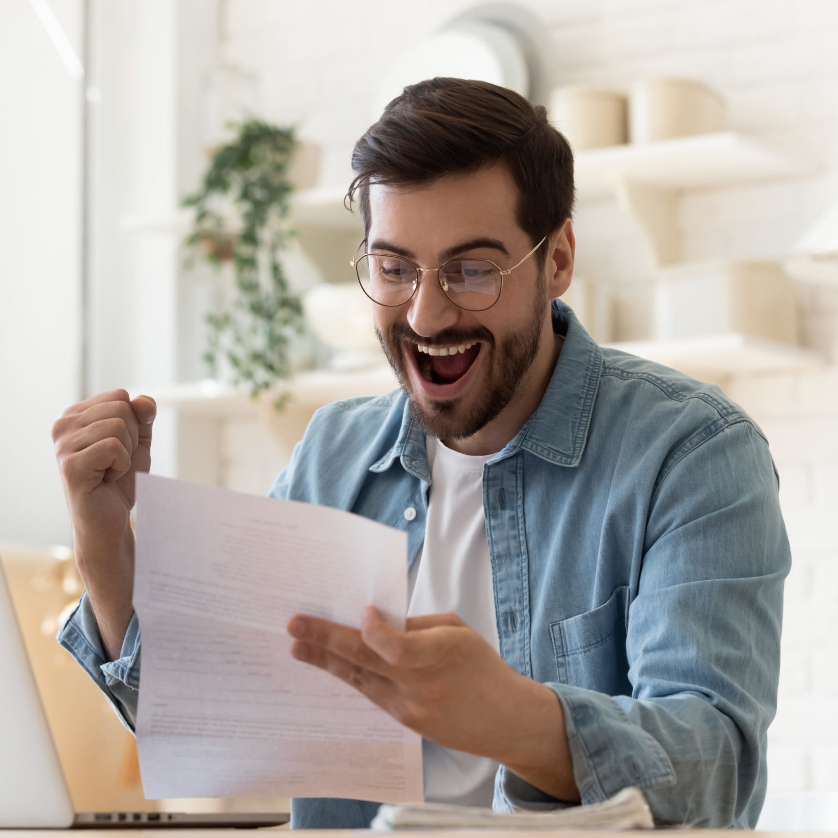 Excited euphoric happy young man holding reading paper postal mail letter amazed overjoyed by good news, got new job celebrate taxes refund receive salary payment loan approval sit at home table